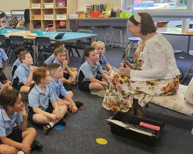 several students sitting on carpet in classroom listening to a teacher speaking